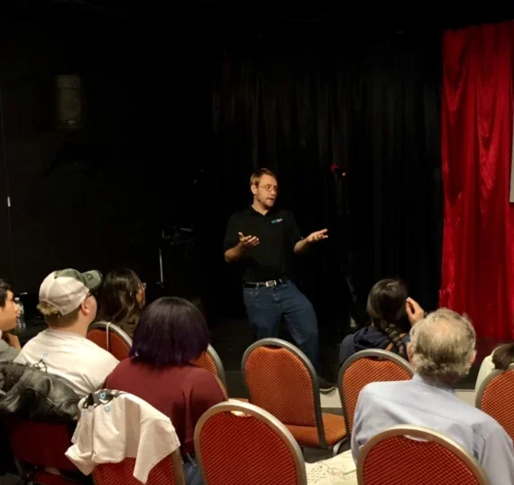 Nick Gausling wearing a collared black shirt and blue jeans, giving a speech to a diverse audience circa 2017-2018